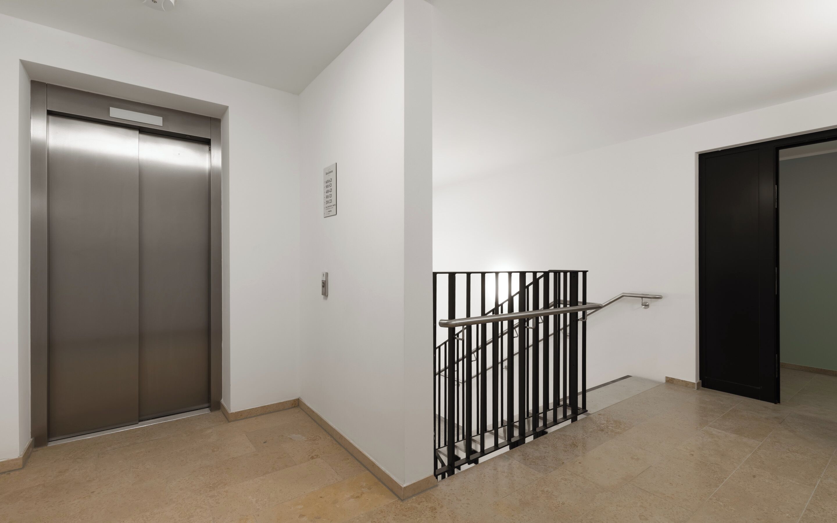 A modern elevator with a metallic finish, a black metal handrail, and a white wall with a door to the right.