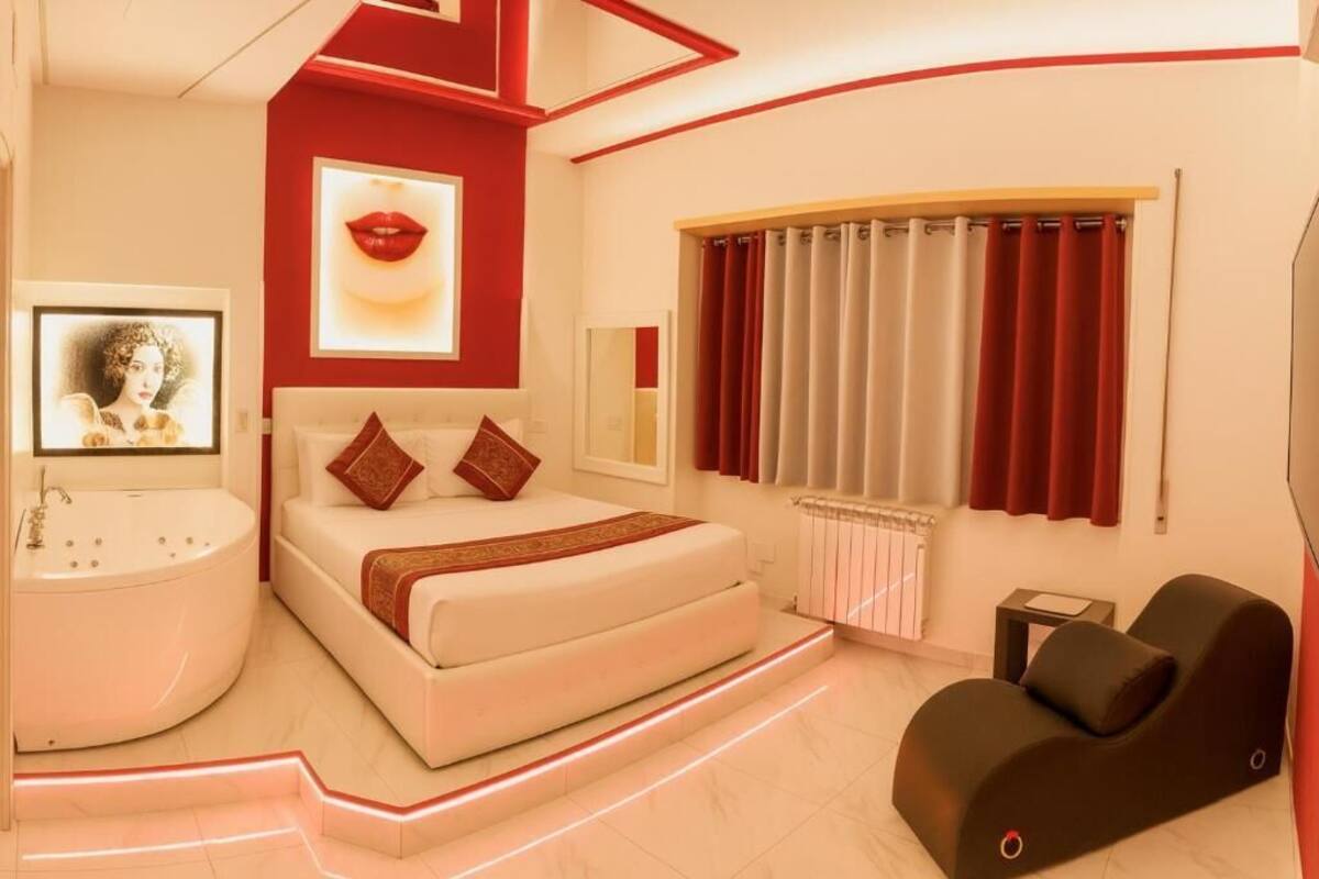 A modern bedroom with a white bed, red accents, a large window with curtains, a bathtub, and a massage chair.