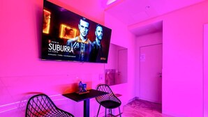 A hotel room with a flat-screen TV mounted on the wall displaying a show, two black chairs with a table in front, and a door to the right. - Bed Cinecittadue (Rome)