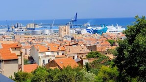 Un paysage urbain côtier avec un port de plaisance, des bâtiments aux toits recouverts de tuiles rouges et un grand navire de croisière bleu et blanc amarré.