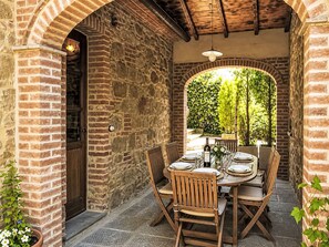 An outdoor patio with a brick wall, wooden table, chairs, and a bottle of wine. - Il Cottage Roccaio by Interhome (Castiglion Fiorentino)