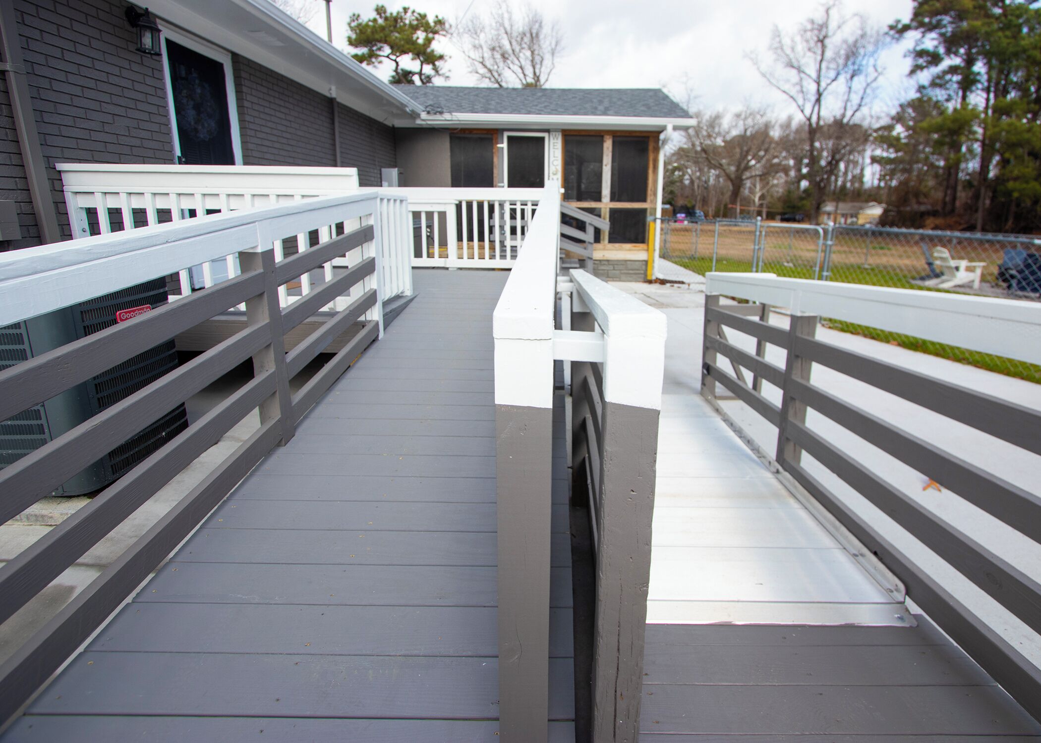 Een overdekt terras met een witte balustrade, dat leidt naar een gebouw met een grijze gevel en een groen dak.