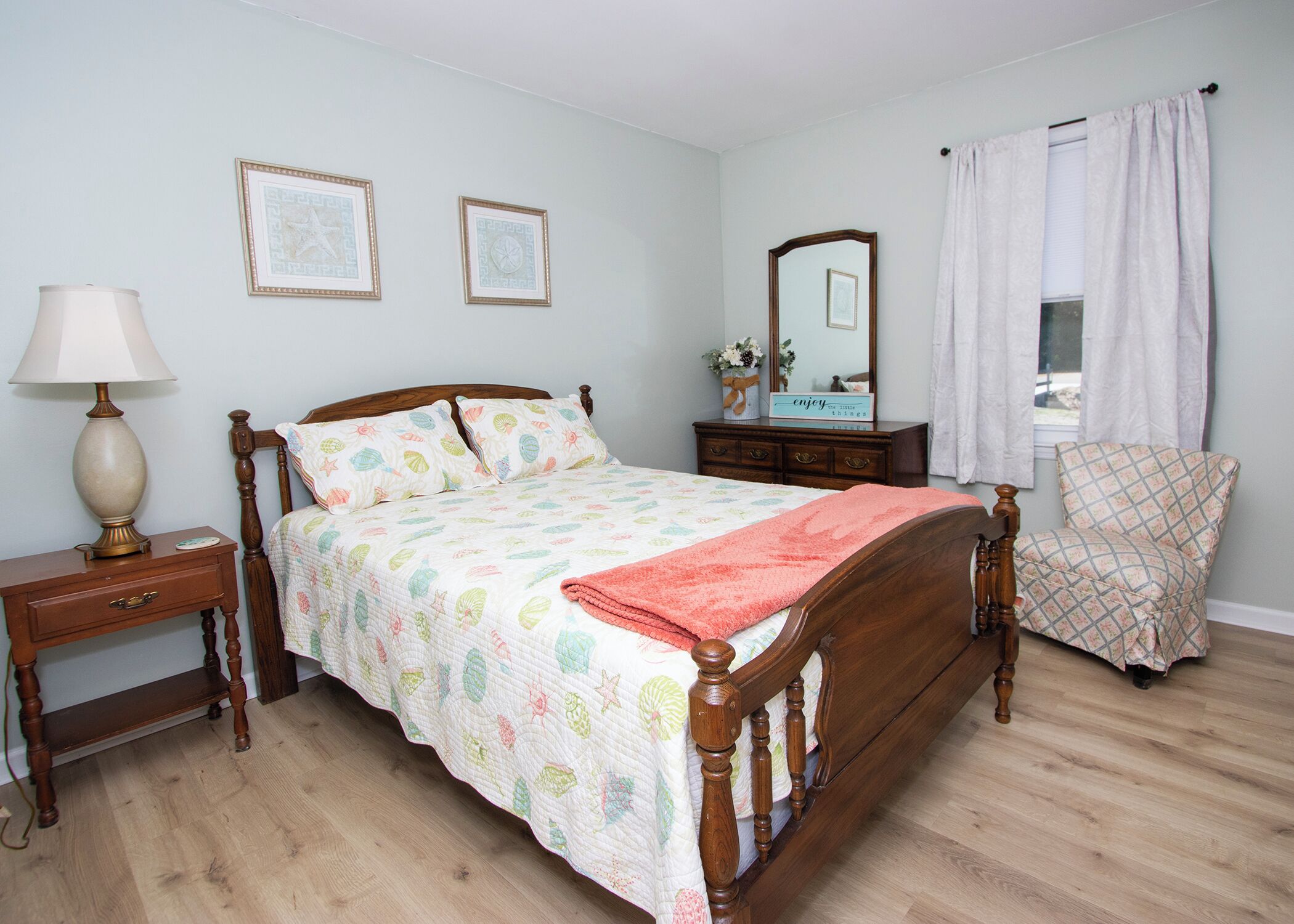 Ein Schlafzimmer mit einem Holzbett, einem Nachttisch mit Lampe, einer Kommode, einem Sessel und zwei gerahmten Bildern an der Wand.