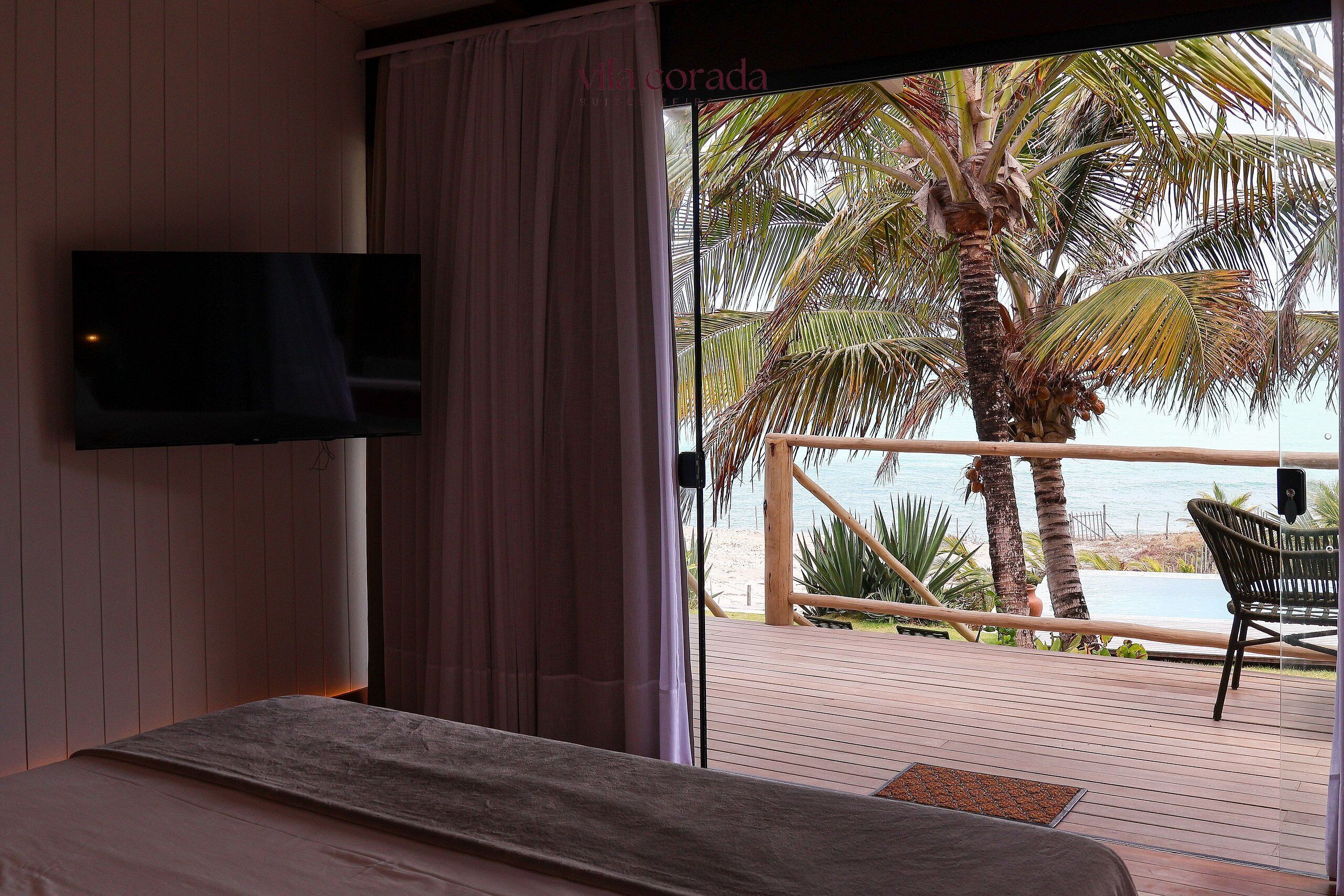 A bedroom with a TV, a bed, a balcony with a view of the beach and palm trees.