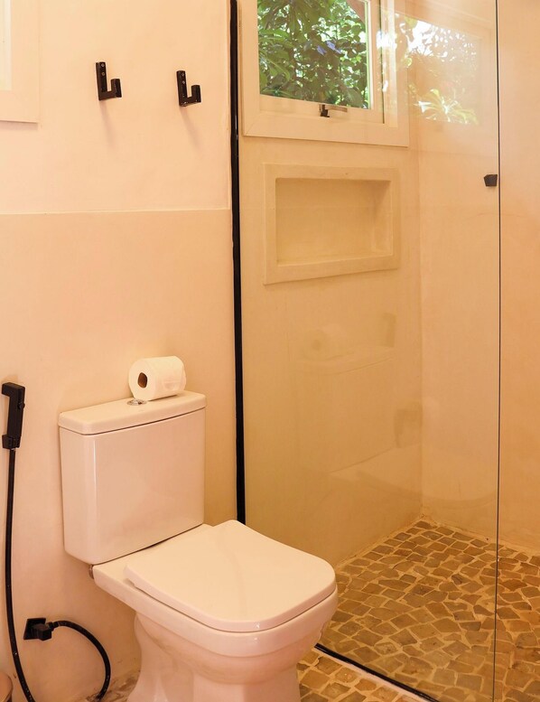 A bathroom with a toilet, a glass shower enclosure, and a window with a view of greenery. - Pousada Vila Corada (Tibau do Sul)