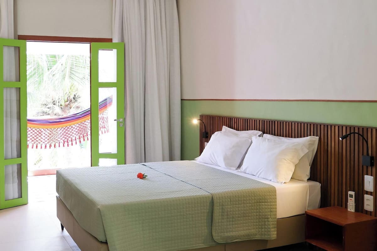 A hotel room with a bed, a nightstand, a TV, and a view of a hammock outside.