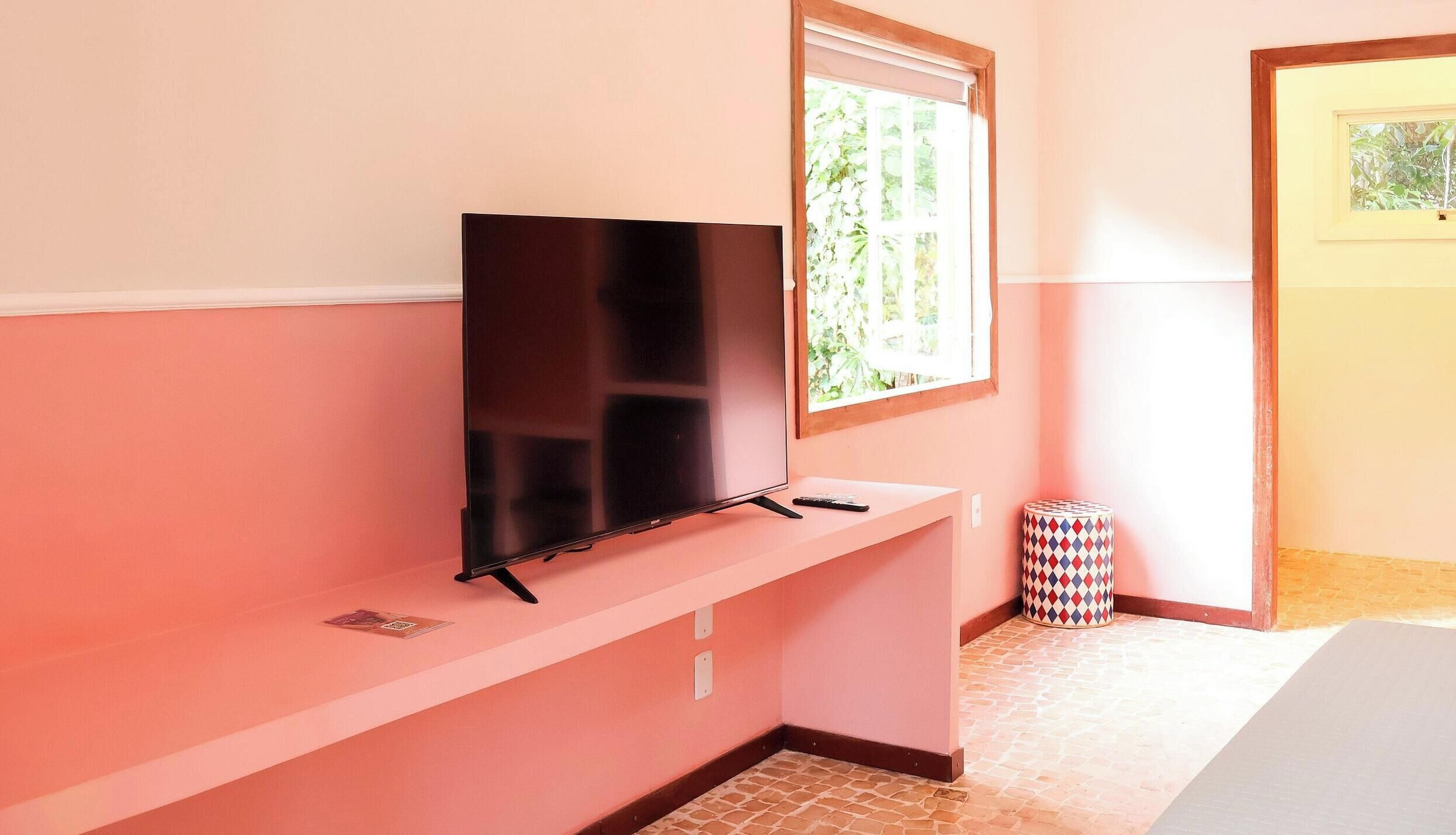 A television on a pink wall, a window with greenery outside, and a patterned trash can.