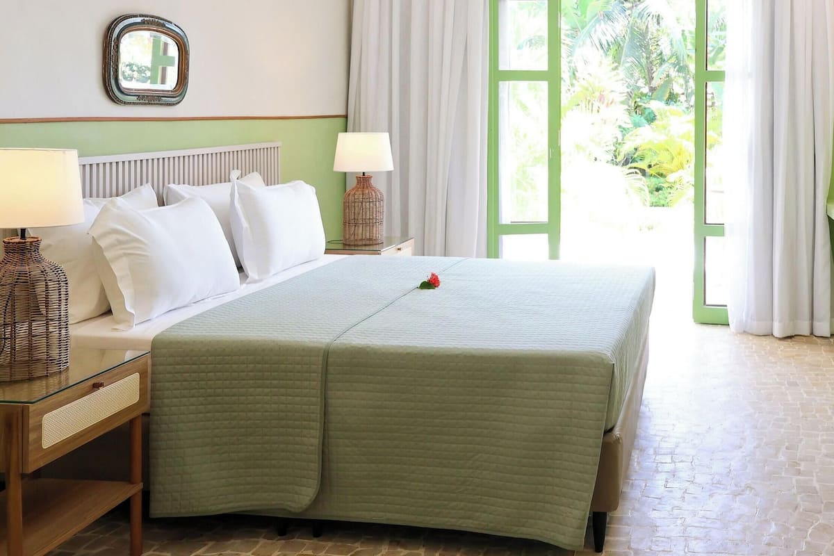 A hotel room with a bed, bedside tables, a lamp, a mirror, and a view of greenery outside.