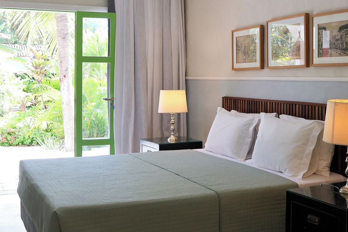 A hotel room with a bed, two bedside lamps, a nightstand, and a view of greenery through a green door.