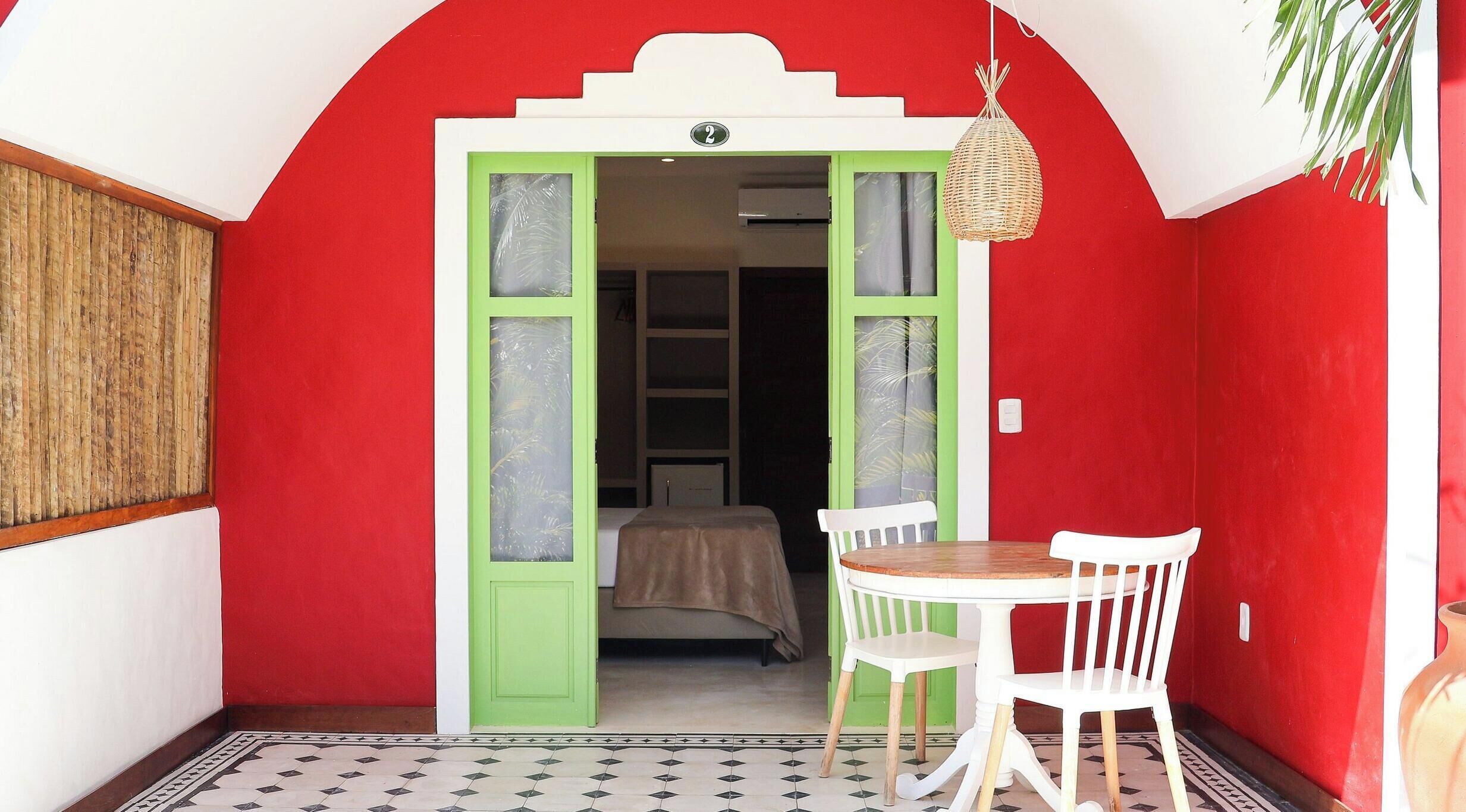 A room with a red wall, a green door, a bed, a table with two chairs, and a hanging light fixture.