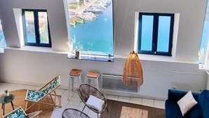 A modern living room with a large window overlooking a marina. There are two windows, a blue sofa, and a wicker chair. - Le Tassigny Duplex Superbe Vue sur les Quai Centre (Sète)