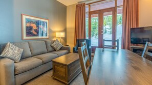 A living room with a sofa, coffee table, and dining table. There is a TV and a painting on the wall.