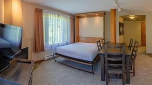 A hotel room with a bed, a desk with a TV, a dining table with chairs, and a window with curtains.