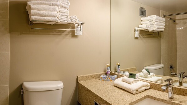 A hotel bathroom with a toilet, sink, and mirror. There are towels stacked on a shelf and a shower area.
