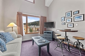 A modern hotel room with a sofa, a table with stools, a TV, and framed pictures on the wall.