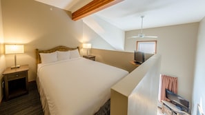 A hotel room with a large bed, two bedside tables with lamps, a ceiling fan, a TV on a stand, and a window with blinds. - Cahilty #308 Hotel Room (Sun Peaks)