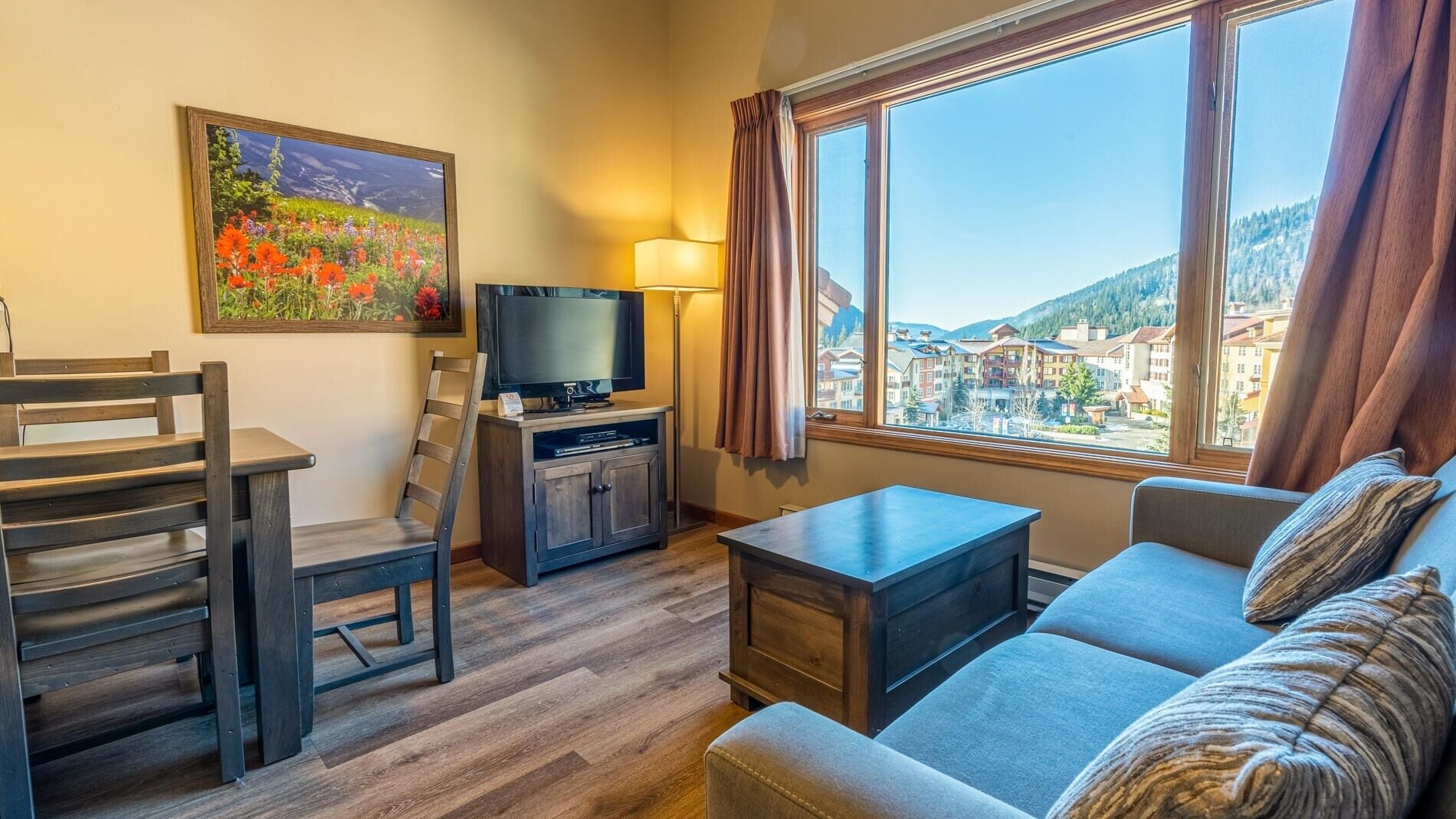 A modern hotel room with a wooden floor, a sofa, a TV, a desk, and a painting of flowers.
