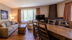 A modern living room with a sofa, a dining table with chairs, a TV, and large windows with curtains. - Cahilty #329 1 Bedroom Hotel Room (Sun Peaks)
