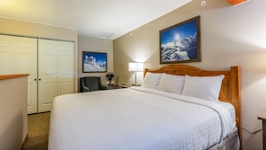 A hotel room with a large bed, a wooden headboard, a desk, a chair, a lamp, and a picture of a snowy mountain.
