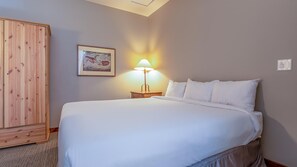 A hotel room with a large bed, a wooden wardrobe, a nightstand with a lamp, and a framed picture on the wall. - Cahilty #436 Hotel Room (Sun Peaks)