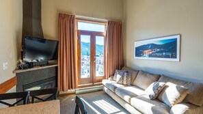 A living room with a television, a sofa, a fireplace, and a large window with curtains. - Cahilty #436 Hotel Room (Sun Peaks)