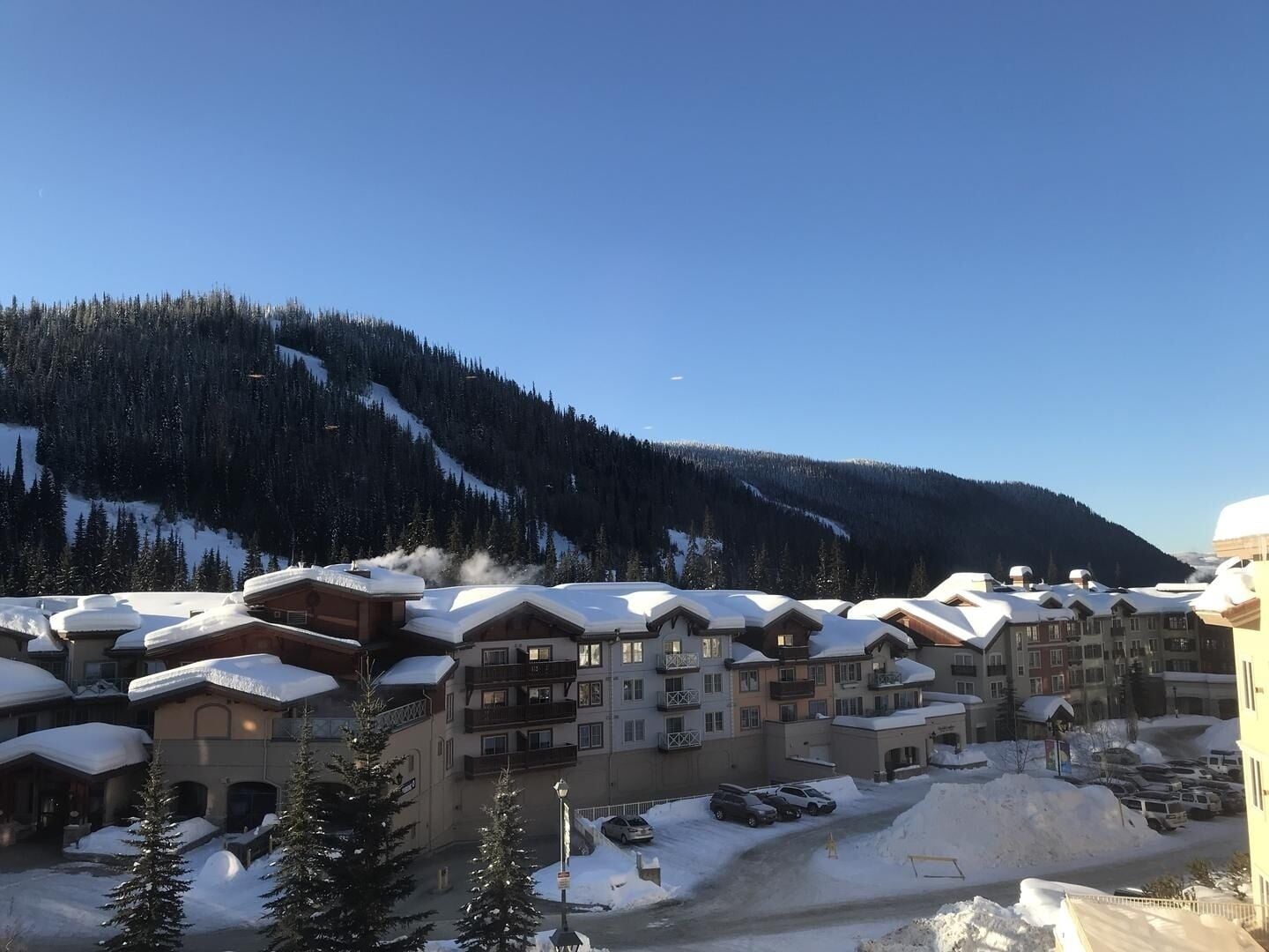 A snow-covered resort with multiple buildings, a ski slope in the background, and a parking area.
