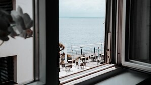 A view through a window of a terrace with tables and chairs, overlooking a body of water. - Apartment "Ella 1" with Lake View, Balcony and Wi-Fi (Friedrichshafen)