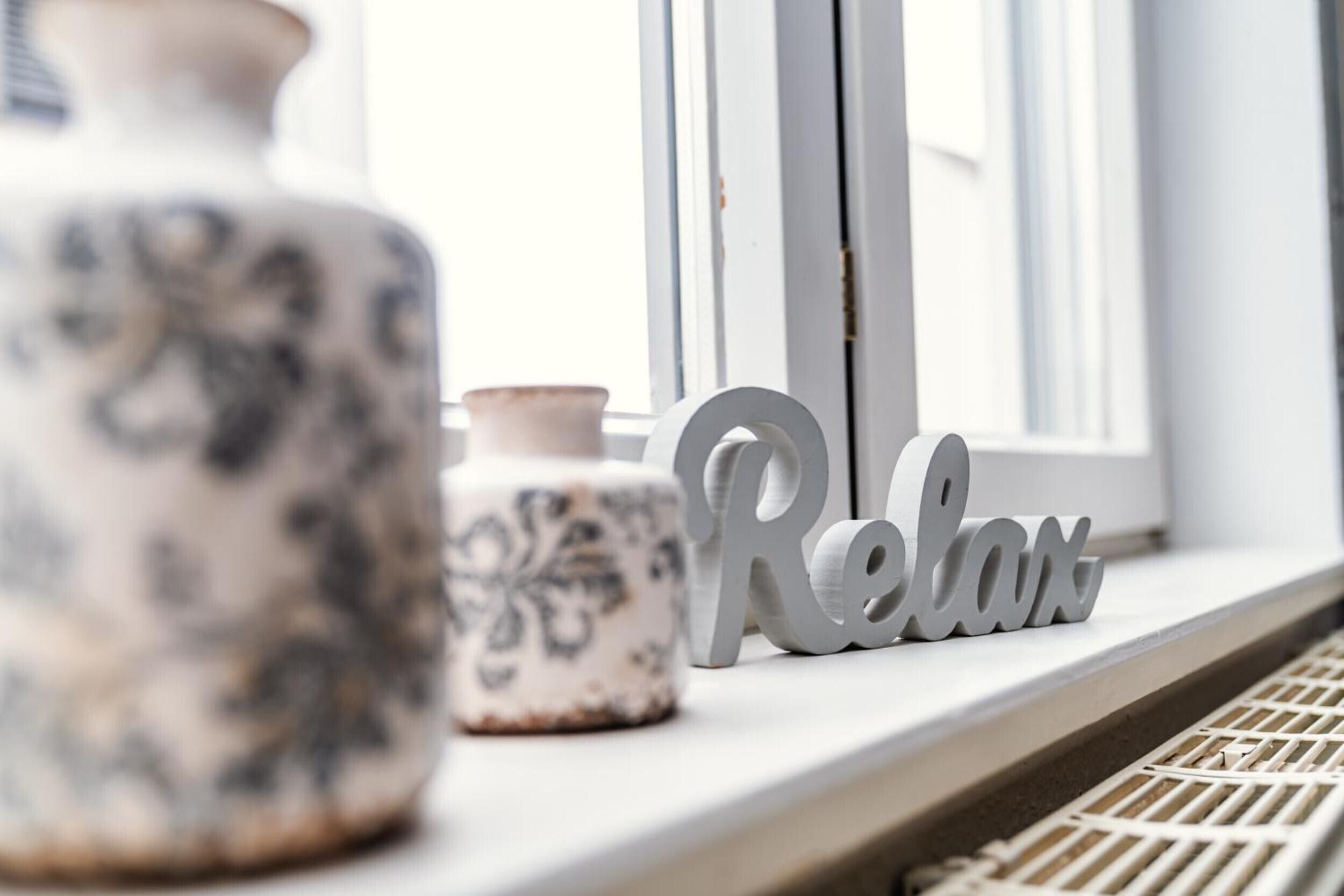 Un alféizar con un jarrón decorativo, una maceta de cerámica más pequeña y un cartel que dice 'Relax'.