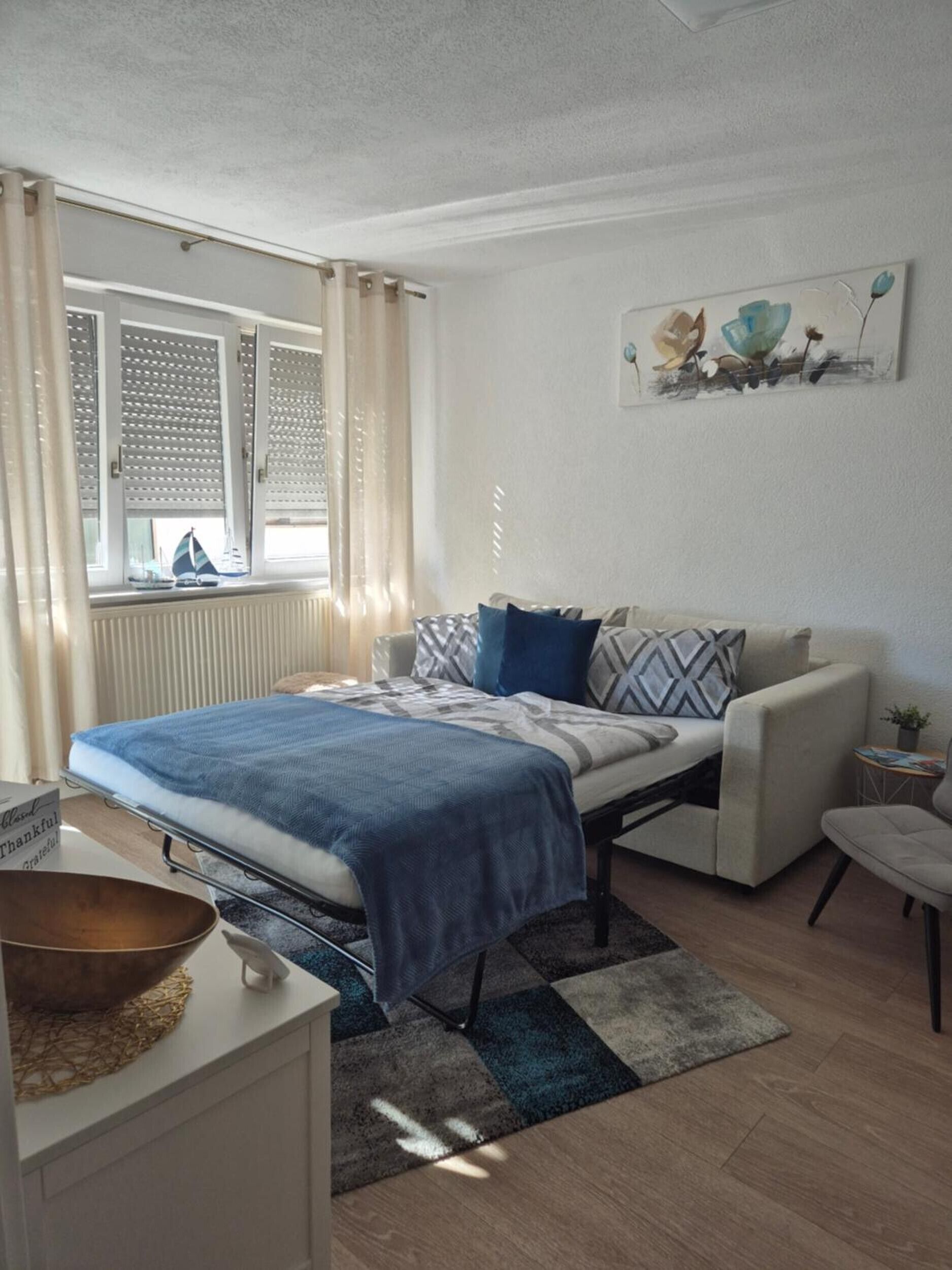 A bedroom with a bed, a sofa, a small table, a window with shutters, and a wall-mounted artwork.