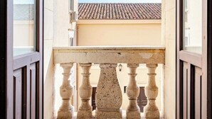 Een balkon met een stenen balustrade, een gewelfde deuropening en uitzicht op een gebouw met een pannendak.