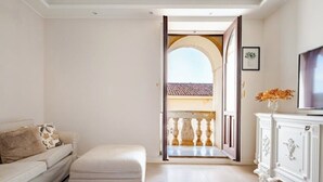 Een kamer met een witte bank, een bijpassend voetenbankje, een witte kast en een televisie. Een open deur leidt naar een balkon met een hekwerk.