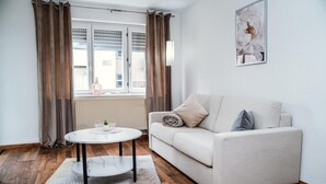 Een woonkamer met een bank, een rond bijzettafeltje, een raam met gordijnen en een schilderij aan de muur.