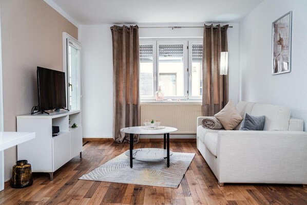 A living room with a sofa, a TV on a stand, a round table, and a window with curtains. - Apartment 'Ella 2' with Lake View, Balcony and Wi-Fi (Friedrichshafen)