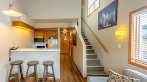 A modern kitchen with wooden cabinets, a built-in microwave, and a refrigerator. There are three stools at the counter and a staircase leading to another level.