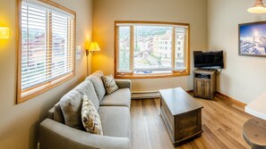 A living room with a sofa, a TV, and a view of buildings through large windows.
