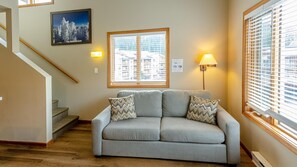 A living room with a sofa, two patterned cushions, a window with blinds, a wall-mounted lamp, a framed picture, and a staircase. - Cahilty #364 Hotel Room (Sun Peaks)