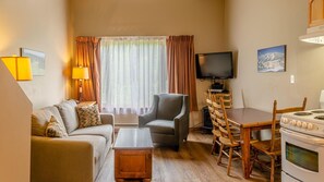 A cozy living room with a sofa, armchair, coffee table, TV, dining table, chairs, and a stove. - Cahilty #427 Hotel Room (Sun Peaks)