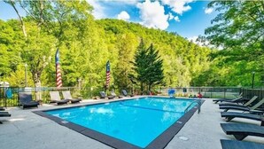 A rectangular swimming pool with lounge chairs and American flags, surrounded by trees and a clear sky. - Creekbend 3 Bedroom Home (Sevierville)
