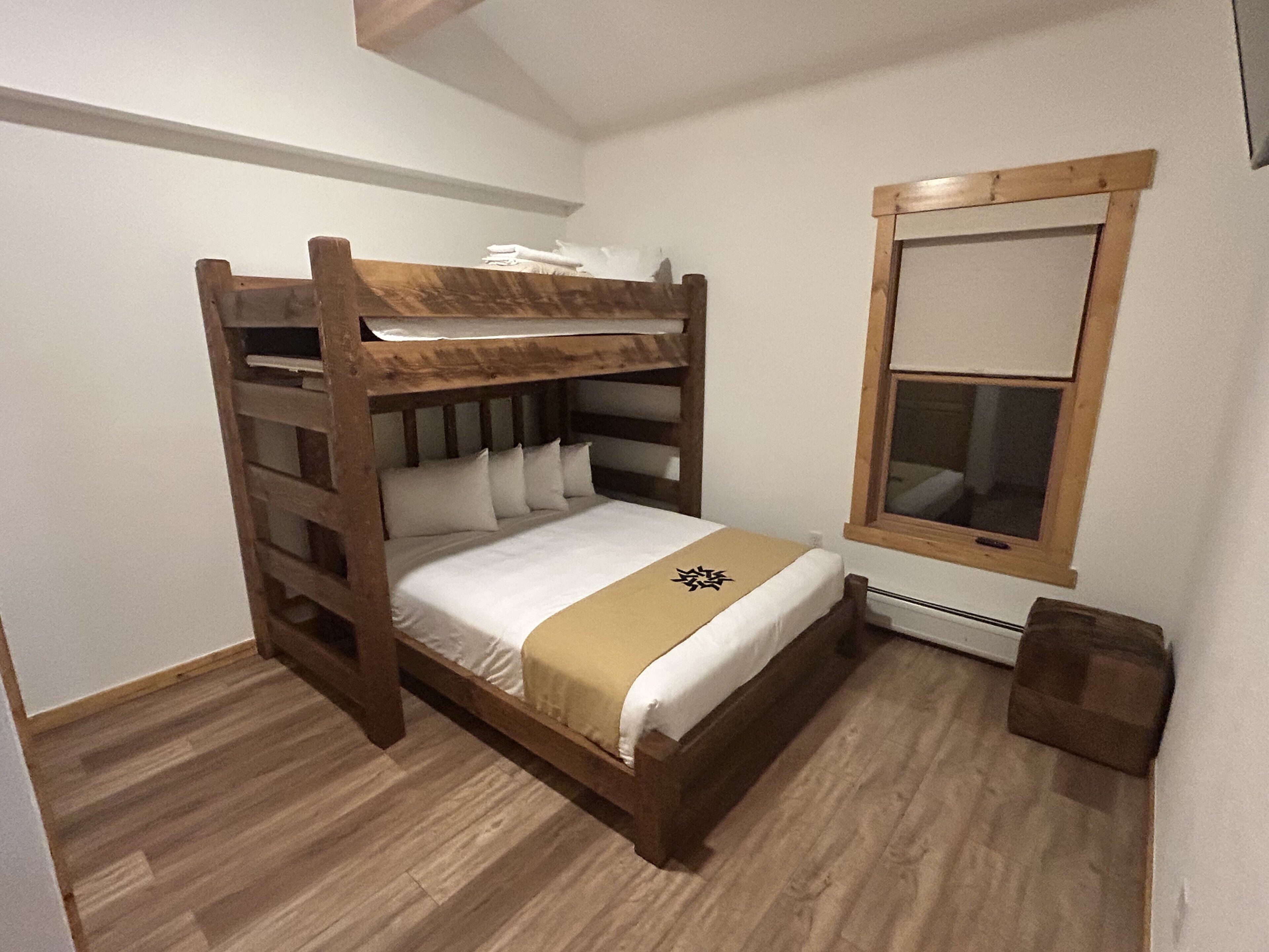 Una cama litera con estructura de madera y ropa de cama blanca. Hay una ventana pequeña con marco de madera en la pared.