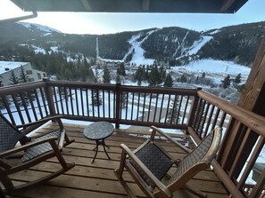 Een houten terras met schommelstoelen en een kleine tafel, met uitzicht op een besneeuwd berglandschap.