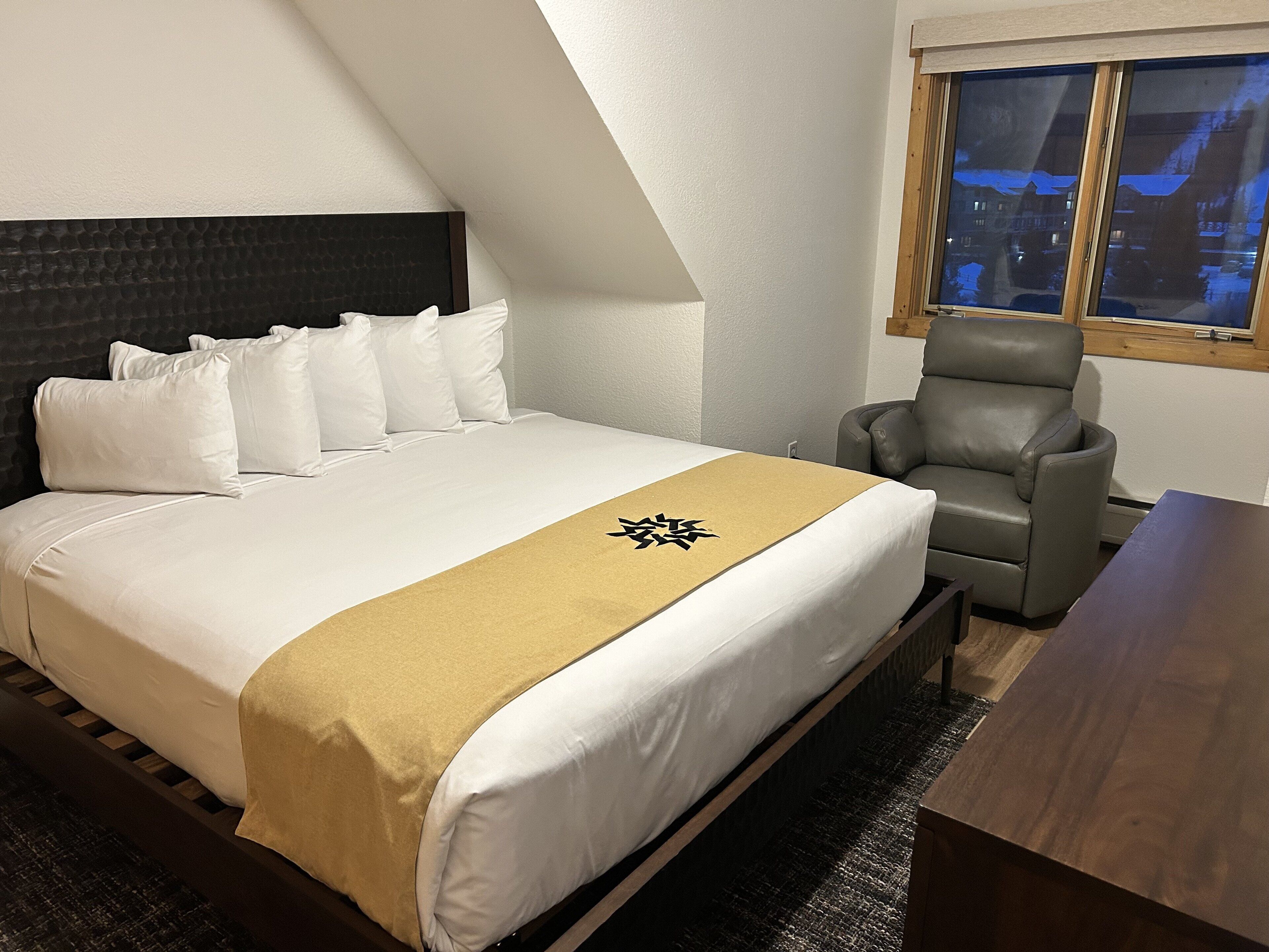 A hotel room with a bed, a grey armchair, a wooden nightstand, and a window with a view of buildings.