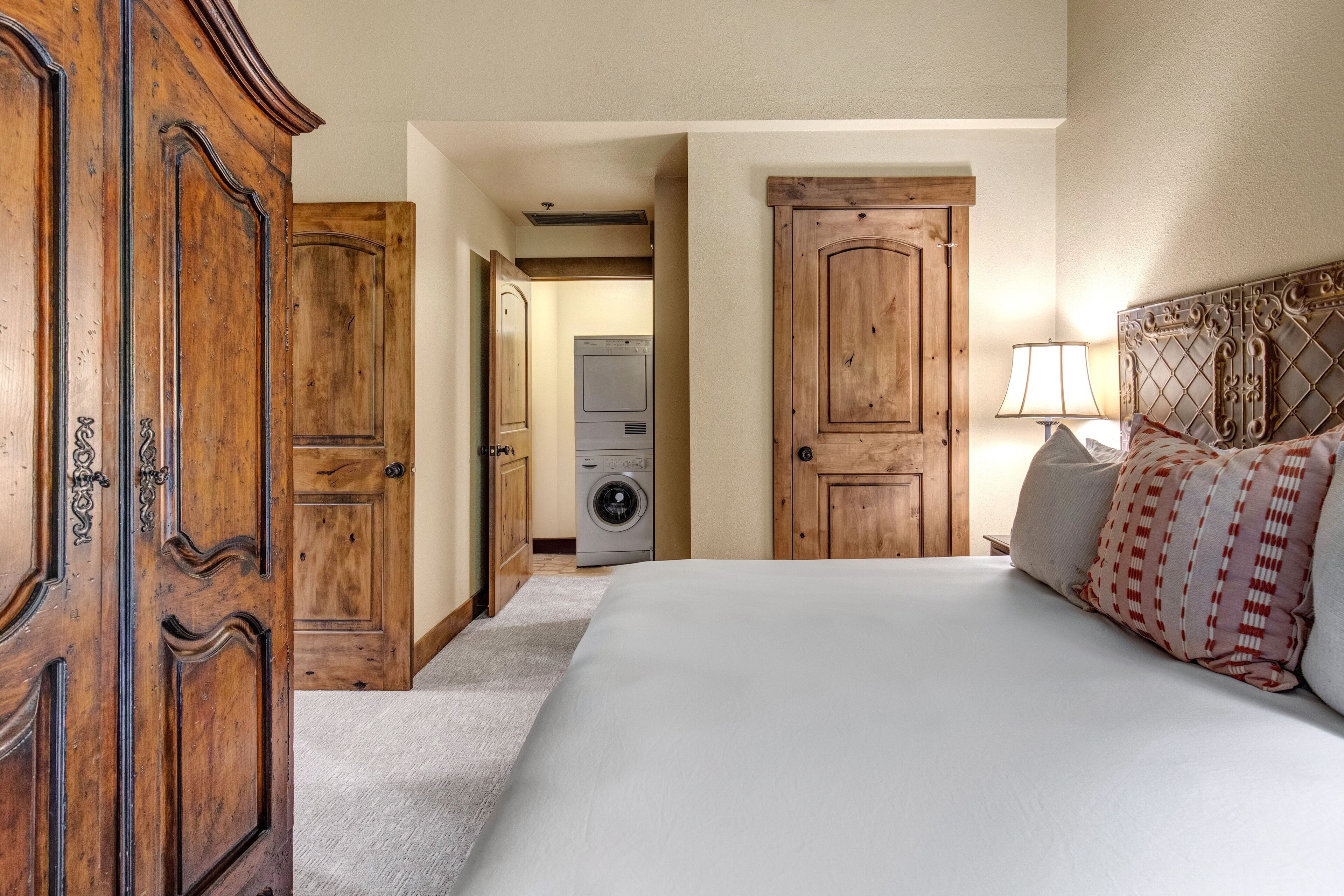 A bedroom with a large bed, wooden wardrobe, and a washing machine in the background.