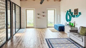 Ruime kamer met houten vloer, hoog plafond met zichtbare balken, en grote schuifdeuren naar een buitenruimte.