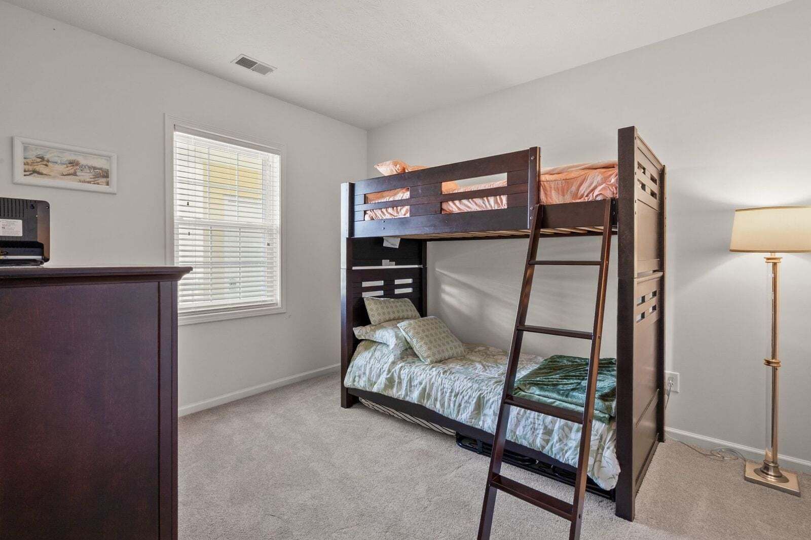 A bunk bed with a ladder, a desk, a lamp, and a window with blinds.