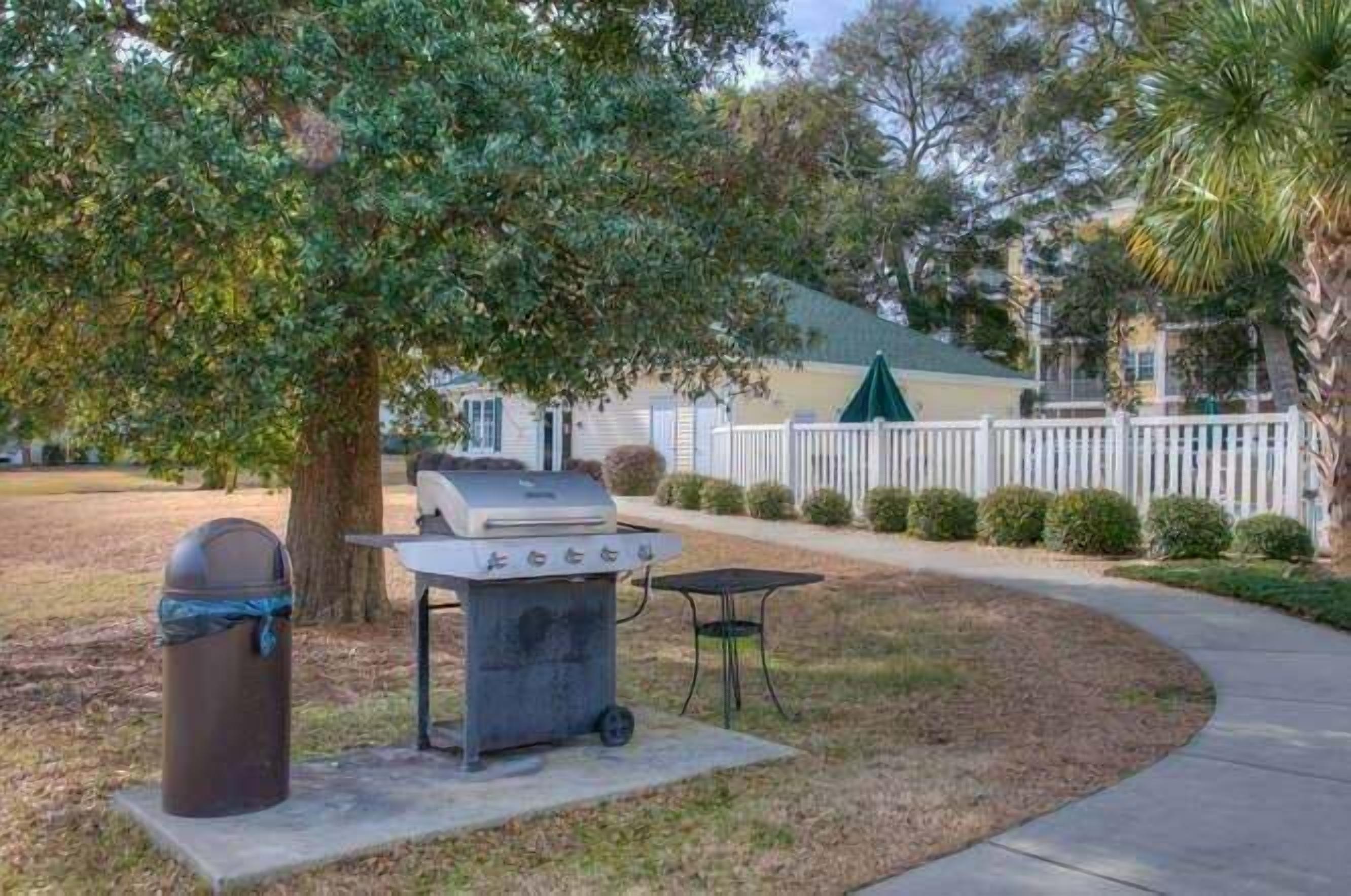 A barbecue grill set up in a grassy area with a tree and a white fence in the background.