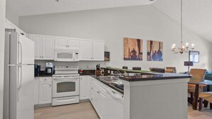 A modern kitchen with white cabinetry, a built-in microwave, a stove, and a dishwasher. There is a dining area with a table and chairs. - Ocean Keyes 2445 3 Bedroom Condo (North Myrtle Beach)