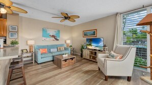 A living room with a blue sofa, a white armchair, a wooden coffee table, and a television. There are ceiling fans, a window with blinds, and a view of palm trees outside. - Big Island Alii Villas 225 1 Bedroom Condo (Kailua-Kona)