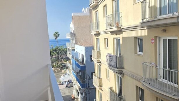 Un edificio de varios pisos con balcones, vista despejada al mar y una calle peatonal abajo.
