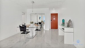 Una habitación moderna y bien iluminada con zona de comedor que incluye una mesa de vidrio y sillas, un mueble blanco con objetos decorativos, y una puerta de madera al fondo.