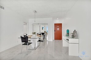 A modern, well-lit room with a dining area featuring a glass table and chairs, a white cabinet with decorative items, and a wooden door in the background. - Balcony-Beachfront Condo+ Gym (Hollywood)